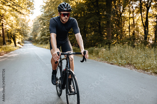Fototapeta Naklejka Na Ścianę i Meble -  Professional cyclist in sport clothing and protective helmet actively riding bike on paved road among green trees. Strong man in mirrored glasses workout on nature.
