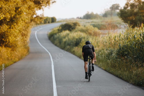 Fototapeta Naklejka Na Ścianę i Meble -  Back view of strong male cyclist with athletic body shape riding bike at the paved road among trees and green bushes in black protective helmet and sportswear. Concept of training  outdoors