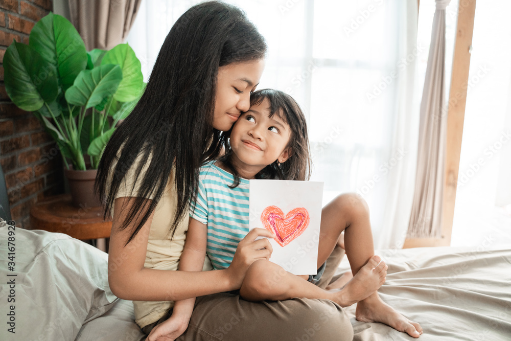 little sister giving surprised love card to her older sister sibling at home little sister giving surprised love card to her older sister sibling at home