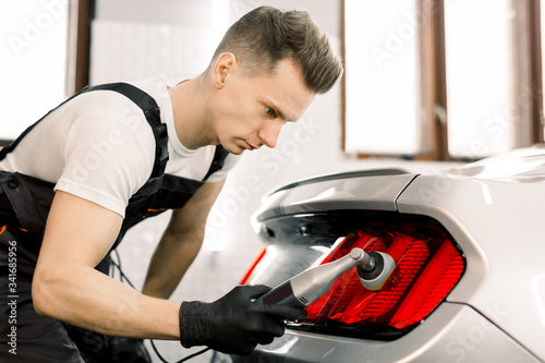 Car detailing series, polishing concept. Professional Caucasian male auto service worker, wearing black protective gloves, waxing and polishing tail light of luxury white car with polish machine