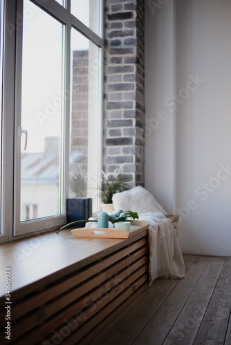 a wide wooden window sill, a large bright window, on a windowsill a wooden tray with a mug, candles. Nearby are books and a green plant in a pot