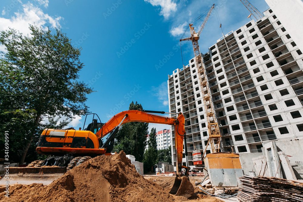orange excavator works on a construction site and a multi-storey residential building under construction on a sunny day