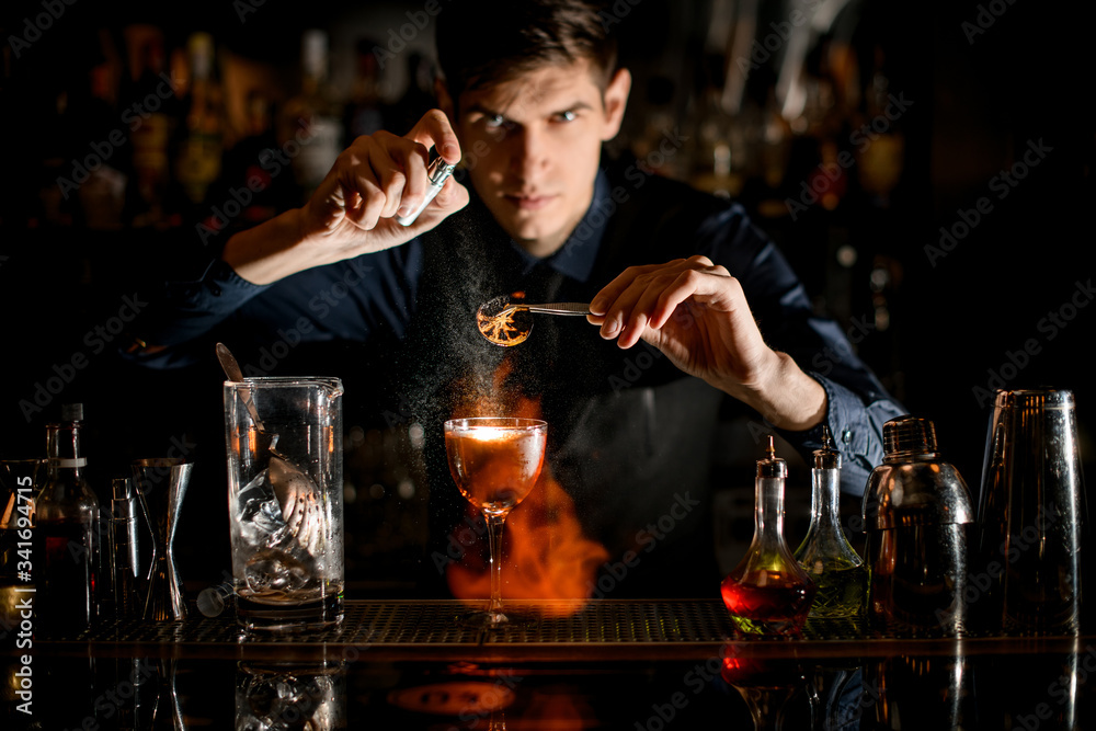 professional bartender holds tweezers with slice of citrus over glass sprinkles and sets it on fire.