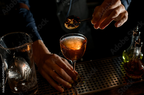 Close-up. Bartender holds tweezers with slice of citrus over glass of drink.