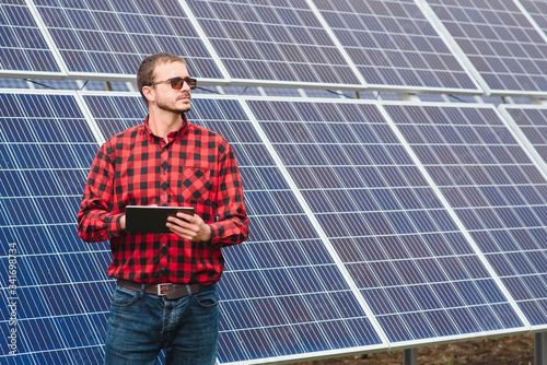 Portrait of man with a tablet in his hands standing near the solar panels station. Green ecological power energy generation. Solar station development concept. Home construction. © Serhii