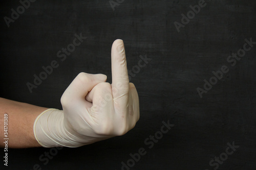 A hand in a white medical disposable rubber latex glove shows the middle finger on a black background, Fuck you. Protective disposable gloves protect against viruses, coronavirus, dangerous bacteria