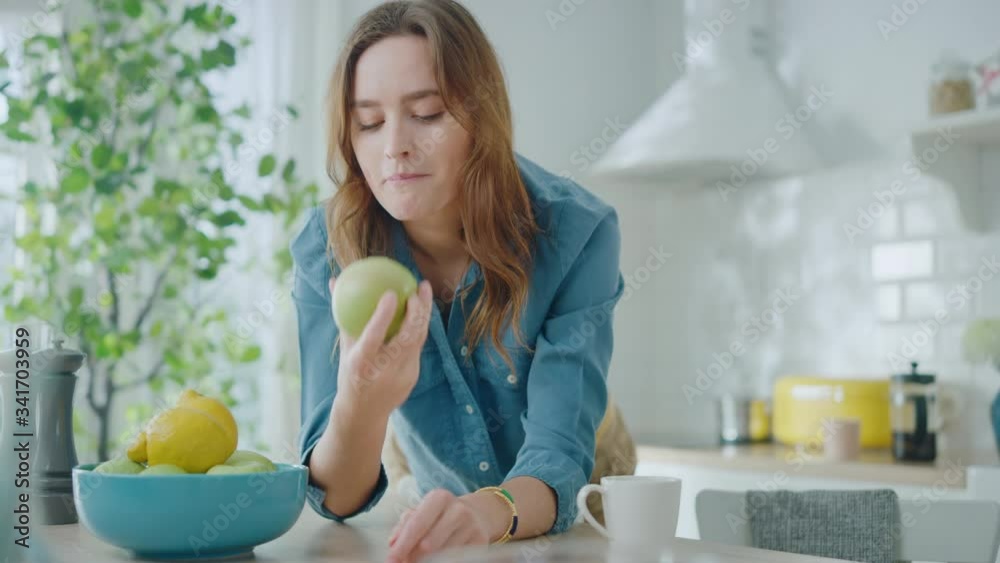 Authentic Beauty Portrait of a Beautiful Brunette Female Eating a Big Green Apple in a Bright Room at Home. Pretty Young Woman Poses for the Camera and Gently Smiles. Healthy Natural Vegetarian. Authentic Beauty Portrait of a Beautiful Brunette Female Eating a Big Green Apple in a Bright Room at Home. Pretty Young Woman Poses for the Camera and Gently Smiles. Healthy Natural Vegetarian.