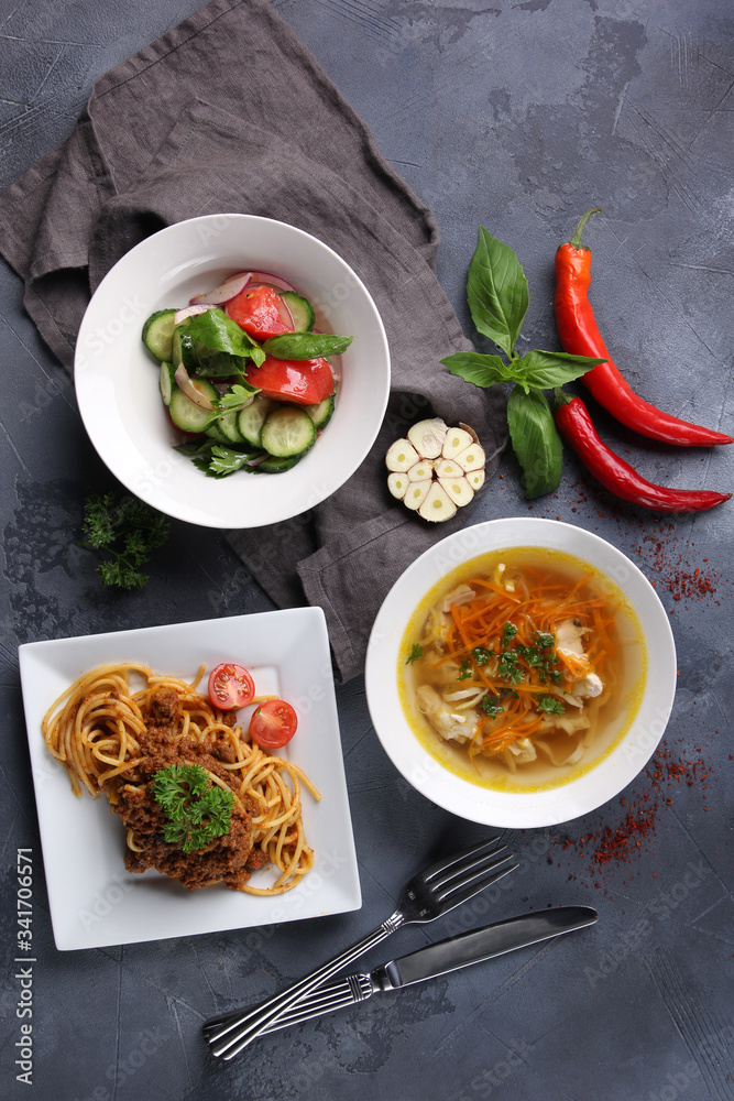 Complex dinner. Fresh vegetable salad, chicken broth and bolognese pasta on white plates on a dark grey background. Fresh herbs, garlic and chili pepper, fork and knife. Top view. Background image