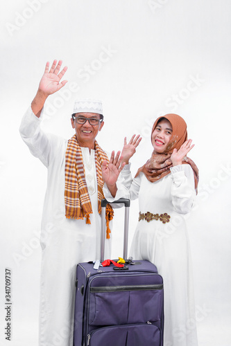 portrait senior asian muslim married couple with travel suitcase. waving their hand to the air and going for ramadan eid mubarak celebration concept isolated over a white