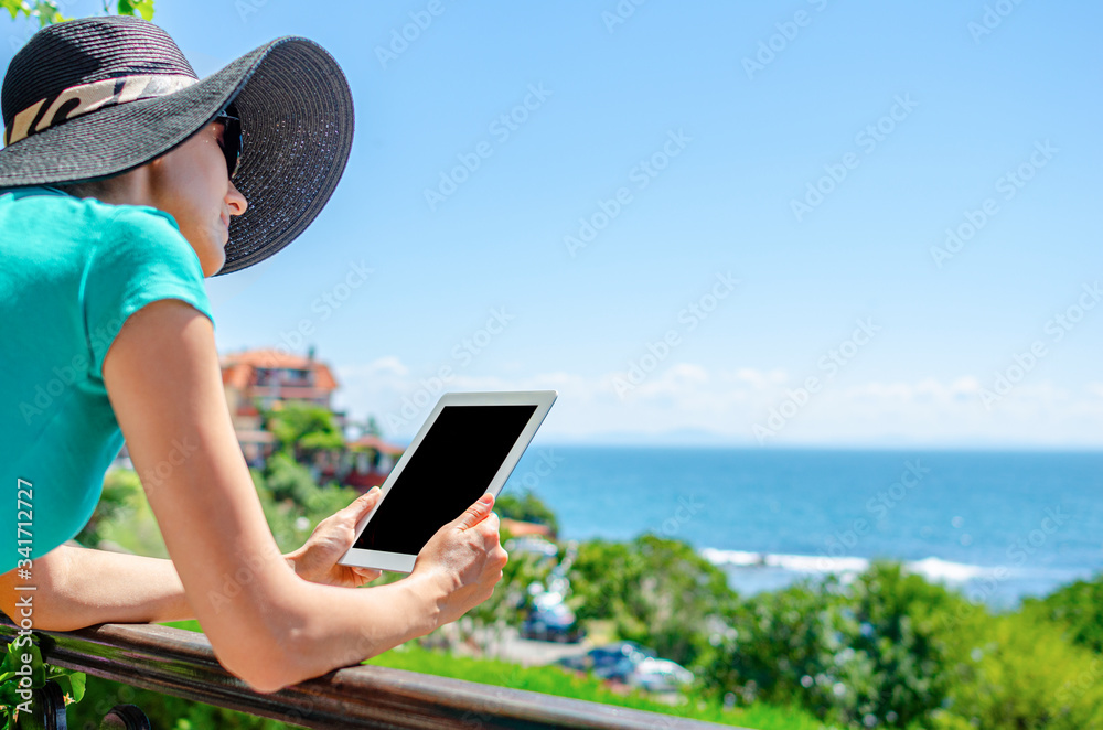 Young woman with a tablet in hands on a terrace on the background of the sea on a warm day.