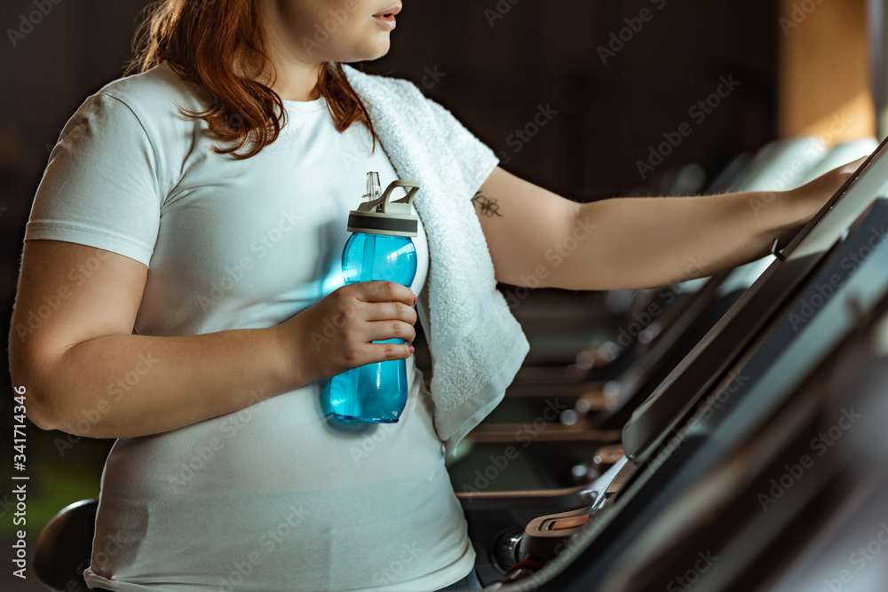 cropped view of overweight girl with towel on shoulder holding sports bottle while standing at treadmill cropped view of overweight girl with towel on shoulder holding sports bottle while standing at treadmill