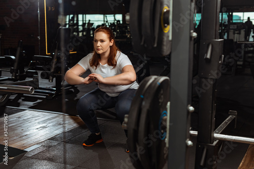 focused overweight girl squatting with clenched hands near fitness machine