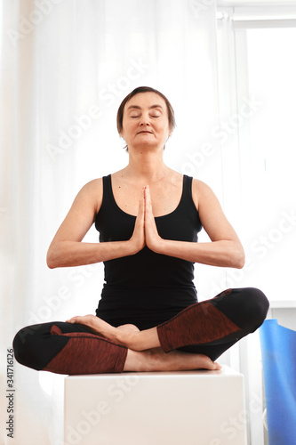 Middle aged woman with hands together close to face in namaste position in the living room. Young looking mature lady during meditation while lockdown epidemic.