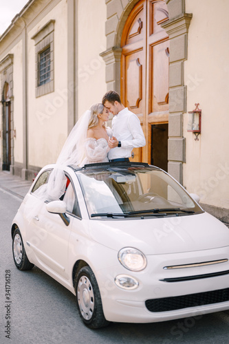 Beautiful bride and groom standing up and leaning forehead to forehead in a convertible in front of Medici Villa of Lilliano Wine Estate, Tuscany, Italy. © Nadtochiy