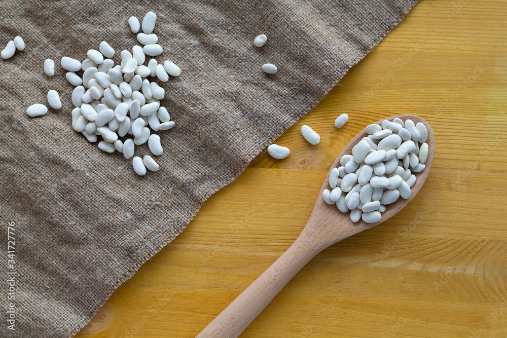 white beans on wooden spoon. wooden spoon with dried white beans and burlap on a wooden table. white beans in spoon lies diagonally with sackcloth on a yellow background. top view. copy space white beans on wooden spoon. wooden spoon with dried white beans and burlap on a wooden table. white beans in spoon lies diagonally with sackcloth on a yellow background. top view. copy space