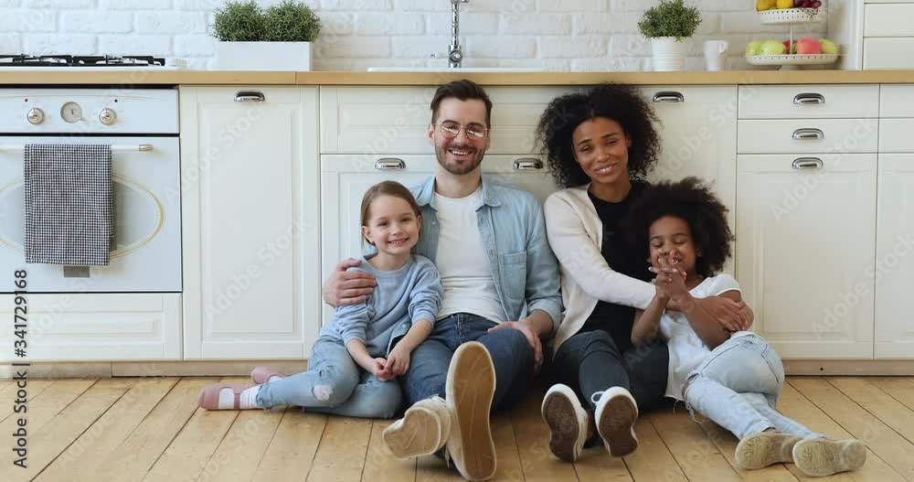 Happy mixed race family young smiling interracial parents mum dad and diverse ethnicity kids daughters sitting on warm heated wooden floor in kitchen at home, hugging, looking at camera. Portrait.