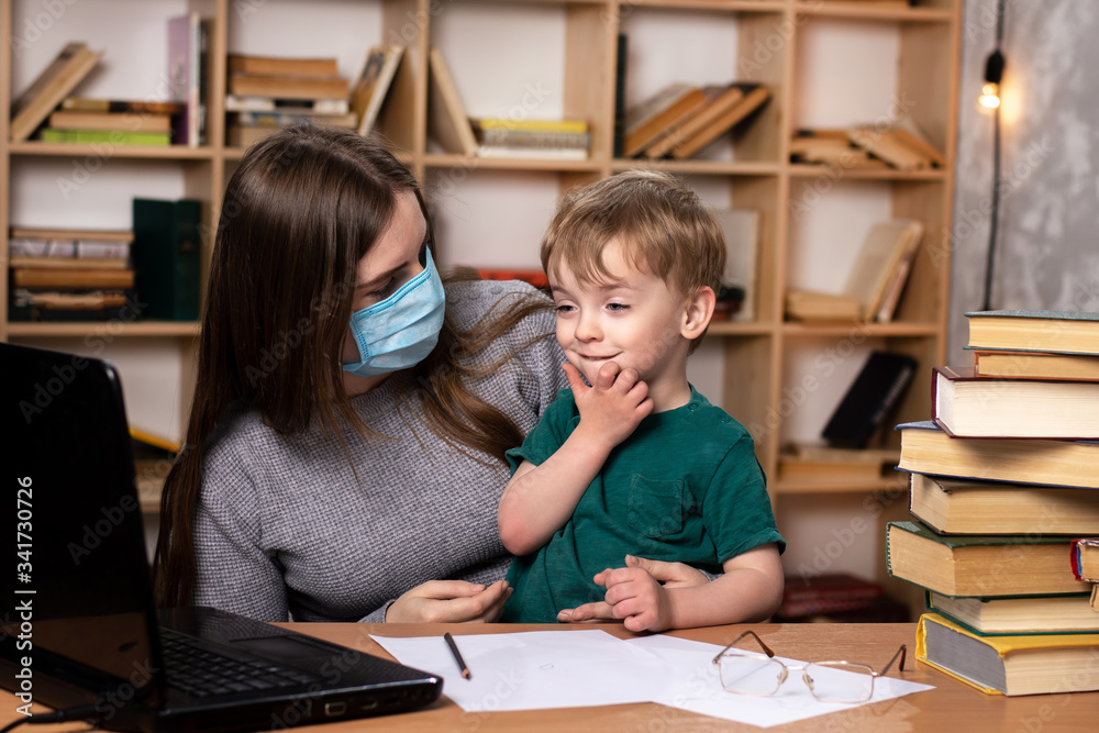 mom in a medical mask with a little boy in her arms at a table with a laptop. family leisure. bookcase on the background. online learning