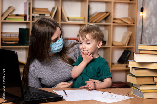 mom in a medical mask with a little boy in her arms at a table with a laptop. family leisure. bookcase on the background. online learning