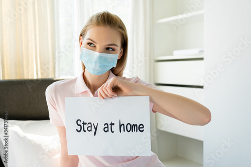 young woman in medical mask holding paper with stay at home lettering in bedroom