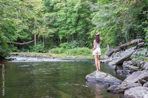 Young girl in white top and cream shorts is standing on big rock and looking on beautiful summer landscape with mountain river and green forest