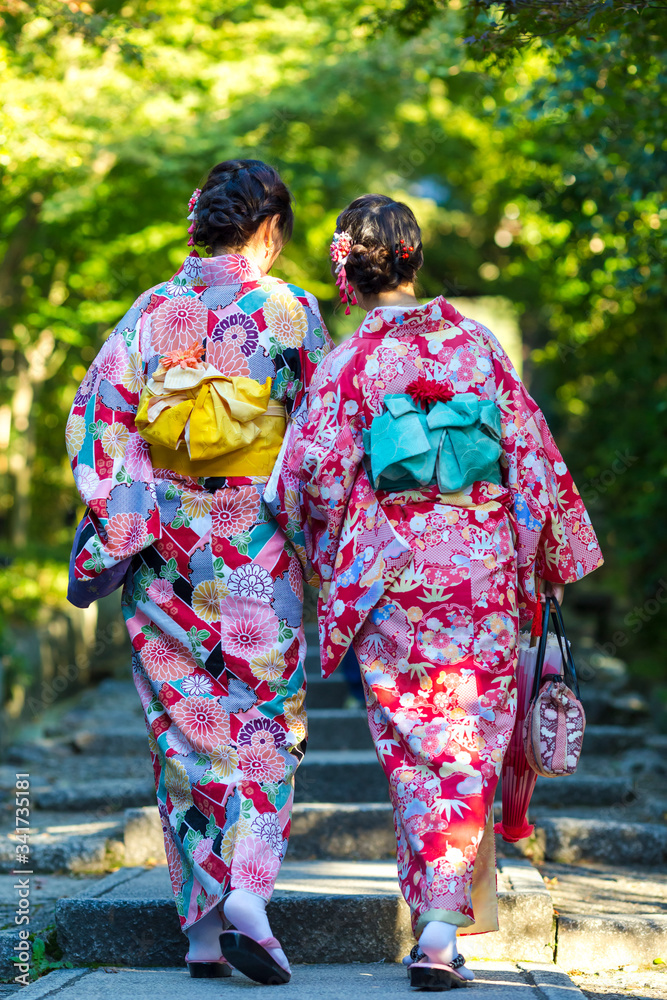 Japan Destination. Two Young Female Geishas in Traditional Japanese Floral Silk Kimono Going Uphill in Green Forest in Kyoto City, Japan. Japan Destination. Two Young Female Geishas in Traditional Japanese Floral Silk Kimono Going Uphill in Green Forest in Kyoto City, Japan.