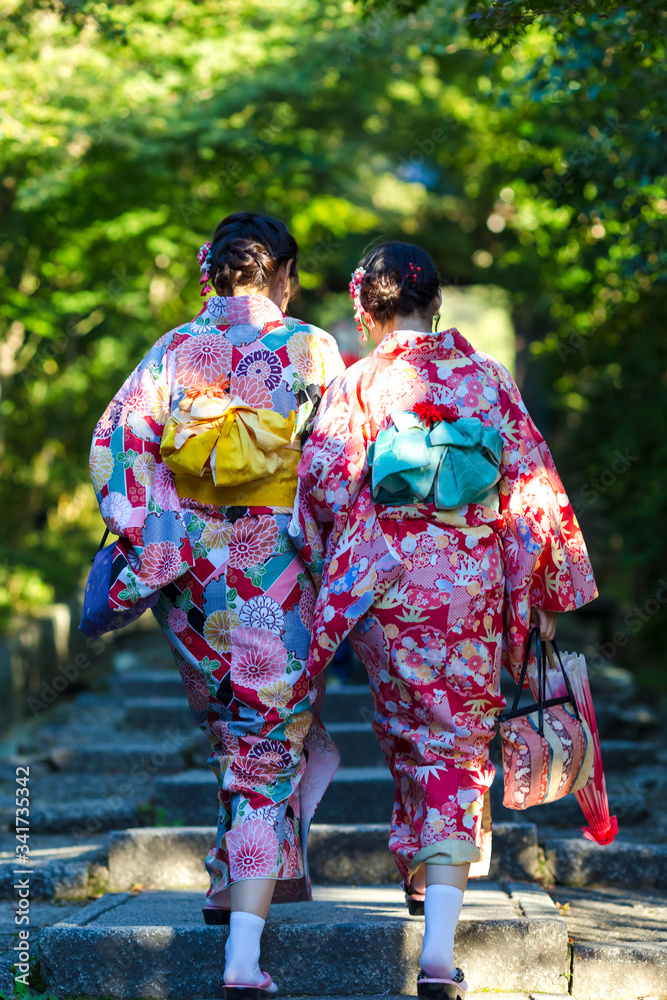 Asian Destination. Two Young Female Geishas in Traditional Japanese Floral Silk Kimono Going Uphill in Green Forest in Kyoto City, Japan. Asian Destination. Two Young Female Geishas in Traditional Japanese Floral Silk Kimono Going Uphill in Green Forest in Kyoto City, Japan.