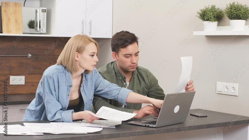 Beautiful lovely young couple reading and analyzing bills sitting at table and using modern laptop. Beautiful lovely young couple reading and analyzing bills sitting at table and using modern laptop.