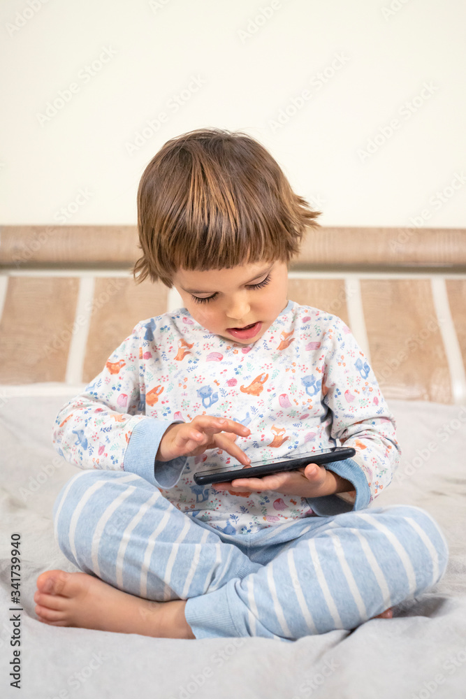 A little boy plays games on the phone. A child sits on a bed in pajamas with a phone