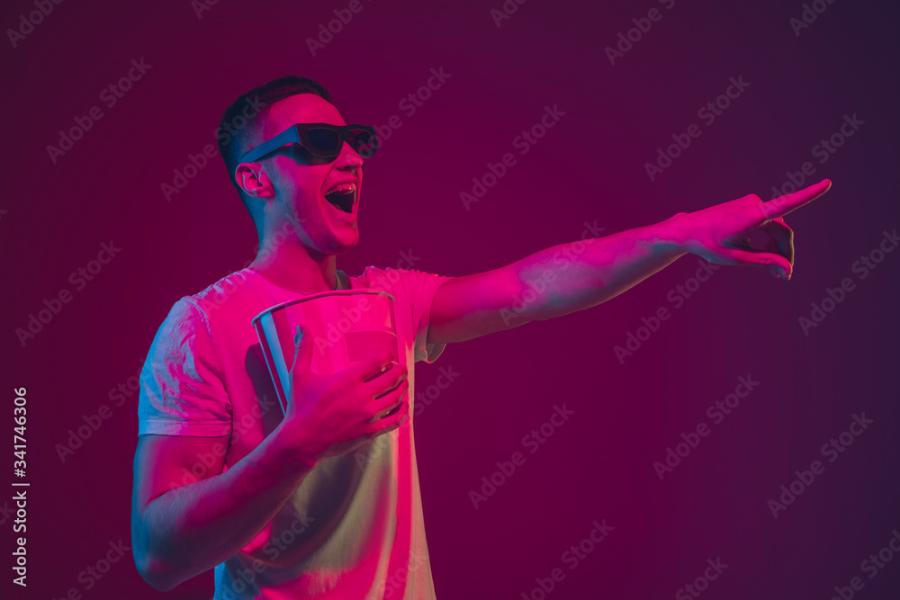 Pointing, holding popcorn. Caucasian man's portrait isolated on pink-purple studio background in neon light. Male model with devices. Concept of human emotions, facial expression, sales, ad. Copyspace Pointing, holding popcorn. Caucasian man's portrait isolated on pink-purple studio background in neon light. Male model with devices. Concept of human emotions, facial expression, sales, ad. Copyspace