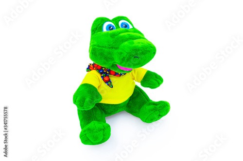 Plush soft, green, children's crocodile toy isolated on a white background with shadow reflection. Front view on the right. The beautiful doll is dressed in a yellow t-shirt and a colorful scarf.