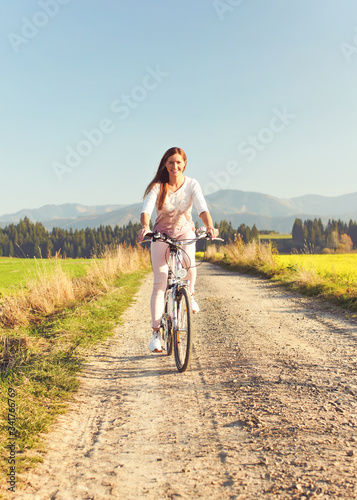 Young woman in summer light clothes rides bicycle on dusty road towards camera, afternoon sun shines to fields and forest in background