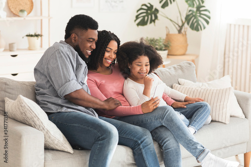 Happy black parents and little daughter relaxing with smartphone at home