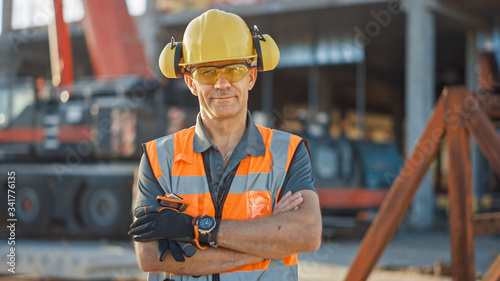 Portrait of Successful Builder / Worker / Contractor Wearing Hard Hat and Safety Vest Standing on a Commercial Building Construction Site, Crosses Arms Confidently. In the Background Crane Machinery photo