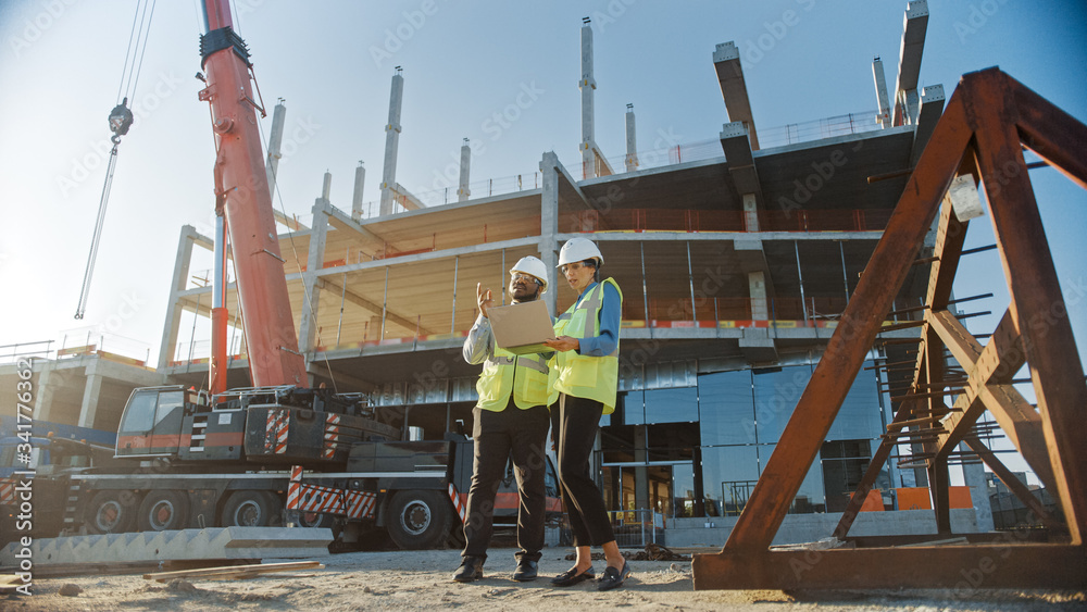 © Gorodenkoff - Two Specialists Inspect Commercial, Industrial Building Construction Site. Real Estate Project with Civil Engineer, Investor Use Laptop. In the Background Crane, Skyscraper Concrete Formwork Frames