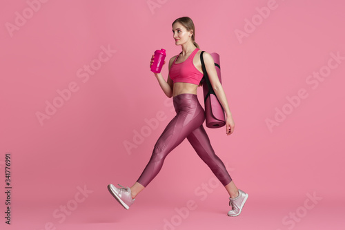 Beautiful young female athlete with bag and water bottle in studio, monochrome pink portrait. Sportive fit caucasian model before training. Body building, healthy lifestyle, beauty and action concept.