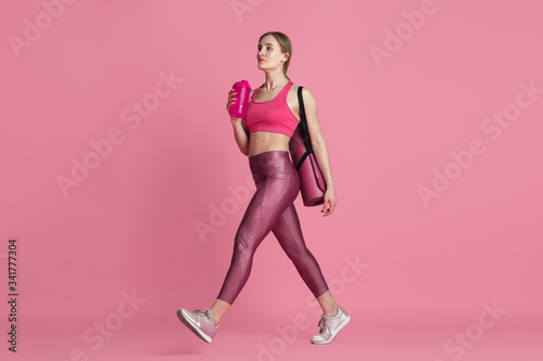 Beautiful young female athlete with bag and water bottle in studio, monochrome pink portrait. Sportive fit caucasian model before training. Body building, healthy lifestyle, beauty and action concept.