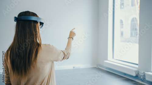 Young Hipster Woman in Holographic Augmented Reality Glasses Standing in Empty Office and Map it. Sunlight Shines Through Big Windows. photo