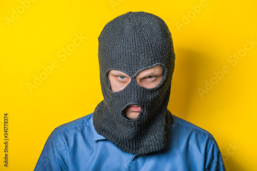man close up thief in a mask and a blue shirt on a yellow background looks slyly to the camera. Mimicry. Gesture. photo Shoot/ evil criminal wearing balaclava © aeroking