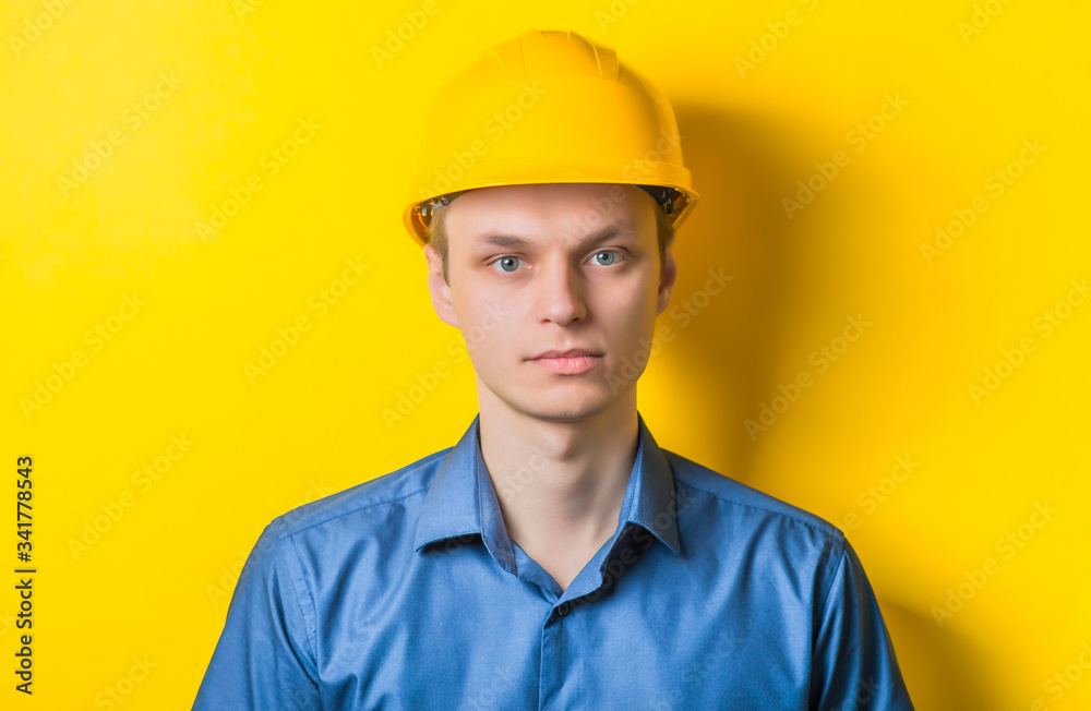 Young man close-up in yellow construction helmet and a blue shirt on a yellow background looking at the camera. Mimicry. Gesture. photo Shoot Young man close-up in yellow construction helmet and a blue shirt on a yellow background looking at the camera. Mimicry. Gesture. photo Shoot