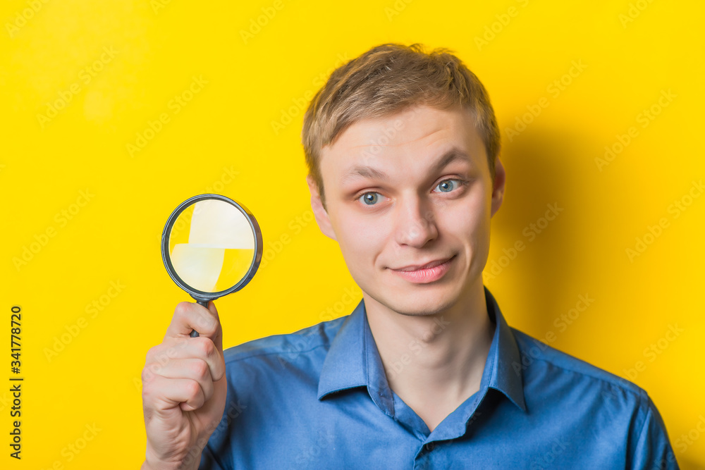 Young man close-up in a blue shirt on a yellow background holding a magnifying glass. Mimicry. Gesture. photo Shoot Young man close-up in a blue shirt on a yellow background holding a magnifying glass. Mimicry. Gesture. photo Shoot