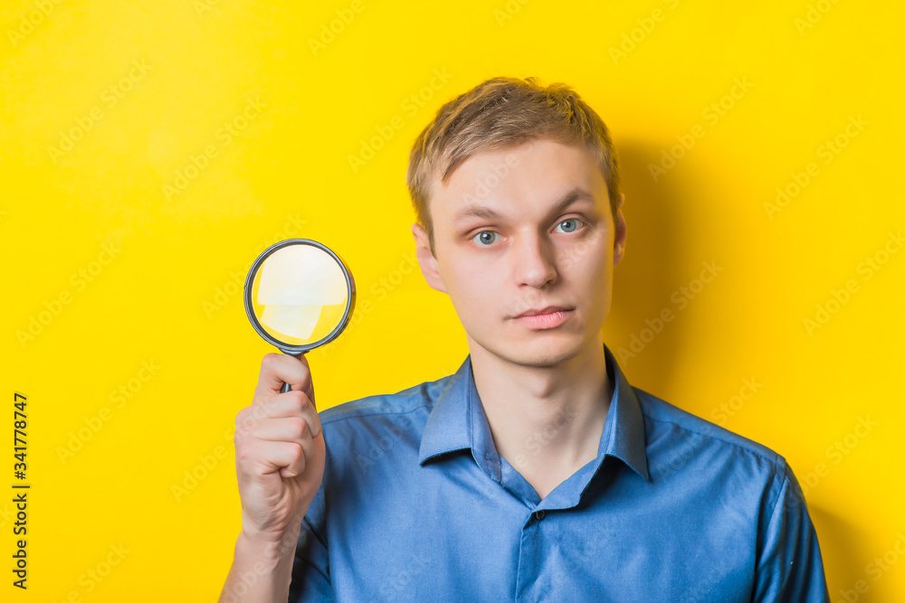 Young man close-up in a blue shirt on a yellow background holding a magnifying glass. Mimicry. Gesture. photo Shoot Young man close-up in a blue shirt on a yellow background holding a magnifying glass. Mimicry. Gesture. photo Shoot