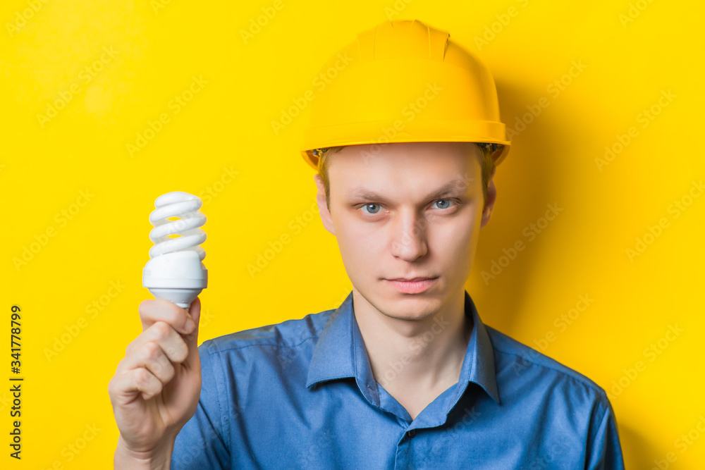 Young man close-up in a blue shirt and construction helmet on a yellow background holding a light bulb energy saving, looking at her. Mimicry. Gesture. photo Shoot