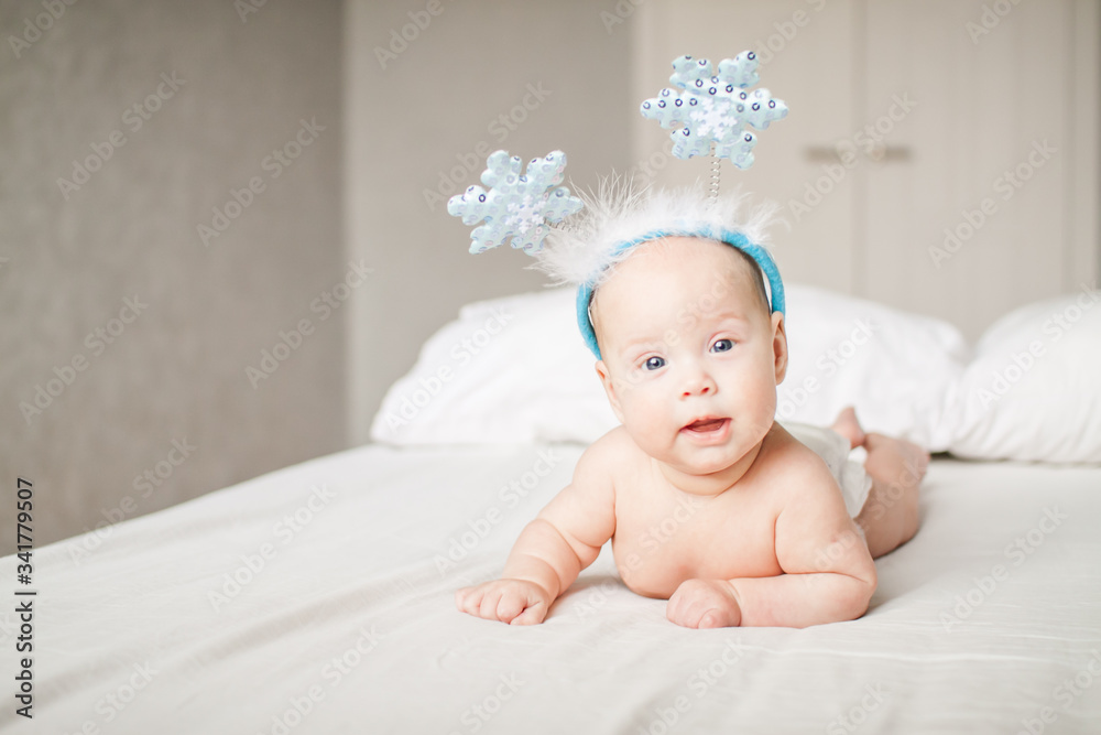 Cute funny infant baby in a rim with horns-snowflakes looking at camera on a white bed. New Year. Cheerful kid. Copyspace Cute funny infant baby in a rim with horns-snowflakes looking at camera on a white bed. New Year. Cheerful kid. Copyspace