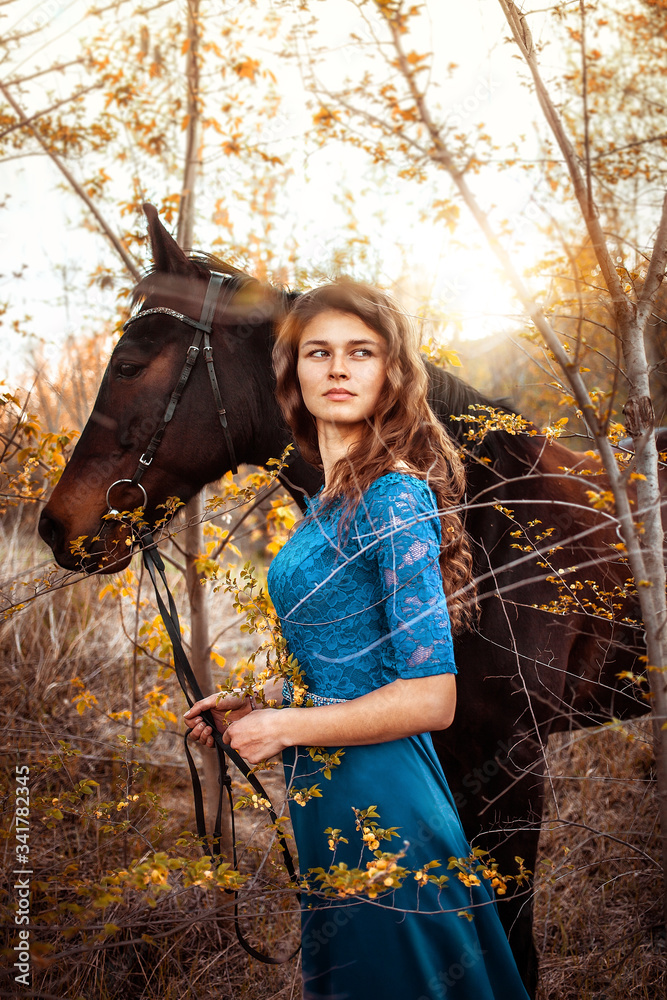 Naklejka premium Beautiful girl with her wavy hair in a blue dress looks into the distance. A bay horse stands behind in a green meadow in the sunset. Magical, fabulous fantasy photography.