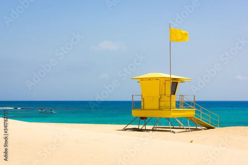 Colorful lifeguard station off duty on empty beach in Corralejo natural park, Fuerteventura. Small fishing boat on turquoise sea with yellow flag on top of baywatch tower on sunny day