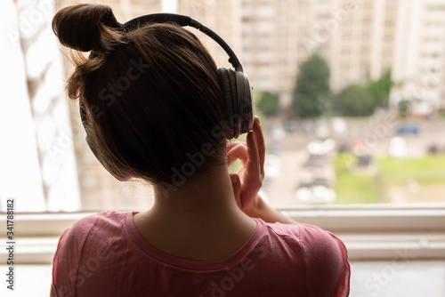 Back view of a lonely young woman listening to music with headphones standing at the window of her apartment. Girl looking on the street