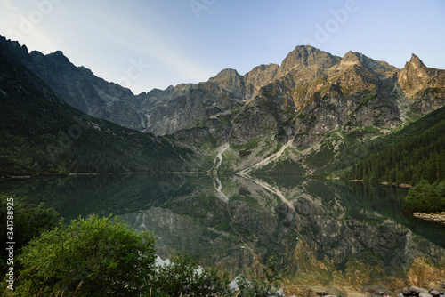 Amazing landscape with high rocks and illuminated peaks, stones in mountain lake(Morskie Oko), reflection at morning sunrise. Concept awakening and harmony with nature. Tatra National Park. Europe.
