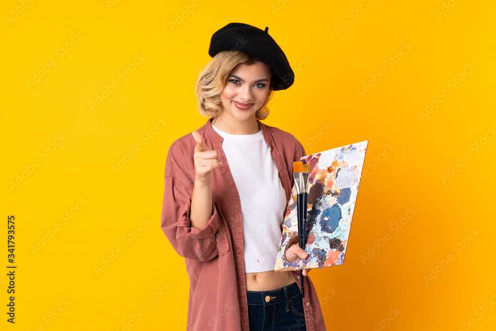Young artist girl holding a palette isolated on yellow background points finger at you