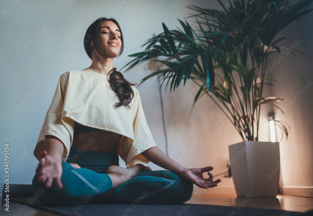 Young happy beautiful woman in cozy cropped sweatshirt and leggings  practicing yoga at home sitting in lotus pose on yoga mat meditating  smiling relaxed with closed eyes Mindfulness meditation concept Stock Photo
