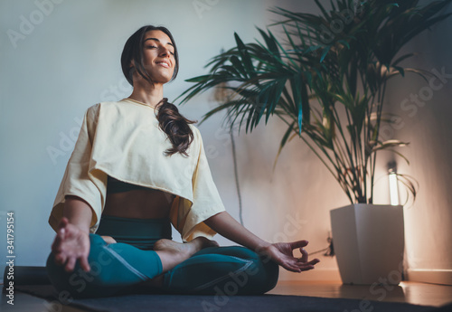 Young happy beautiful woman in cozy cropped sweatshirt and leggings practicing yoga at home sitting in lotus pose on yoga mat meditating smiling relaxed with closed eyes Mindfulness meditation concept